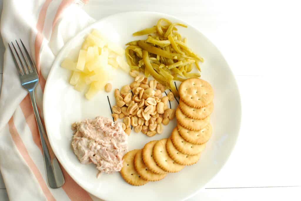 A snack plate with crackers, tuna, fruit, peanuts, and green beans.