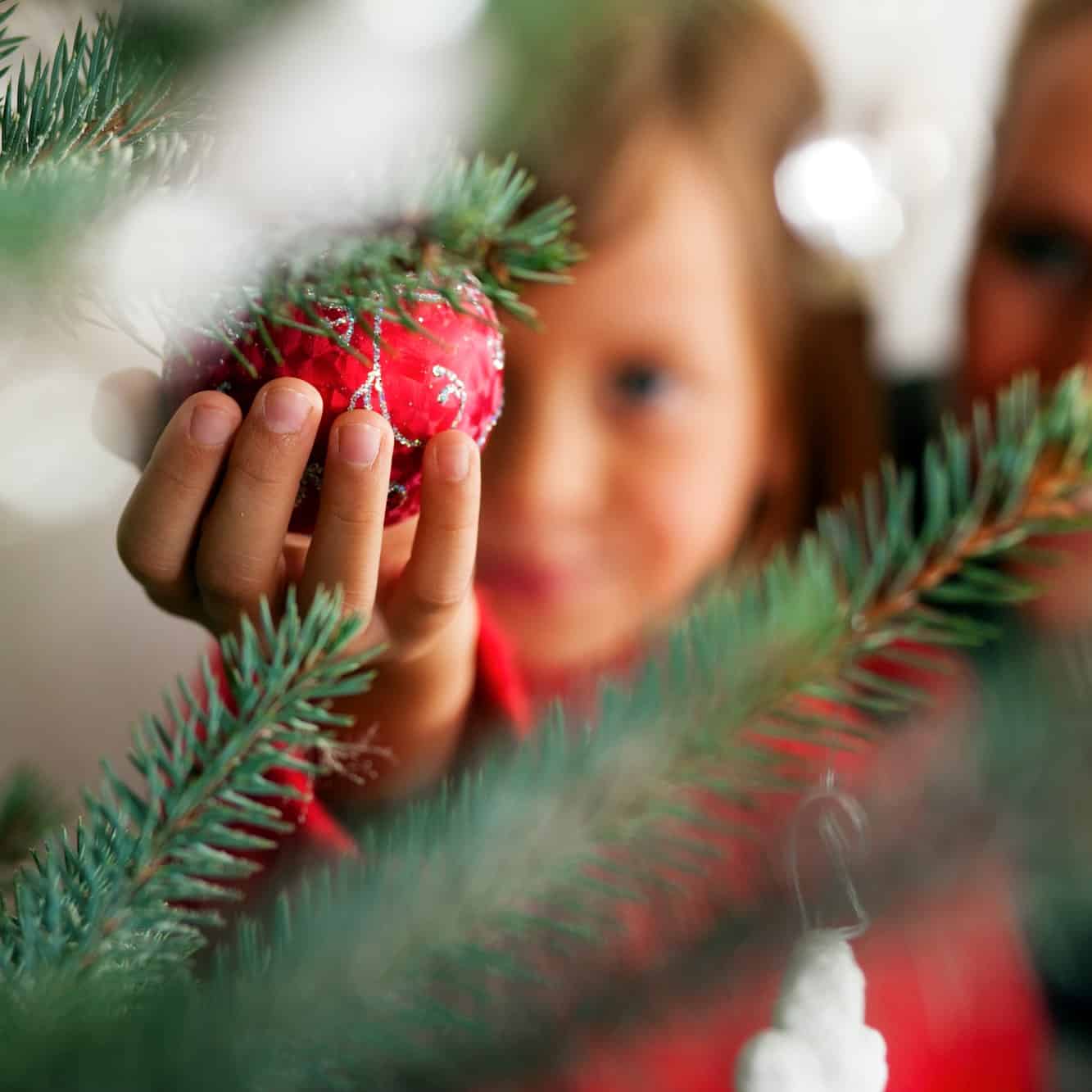 A little girl holding a red ornament near a Christmas tree.
